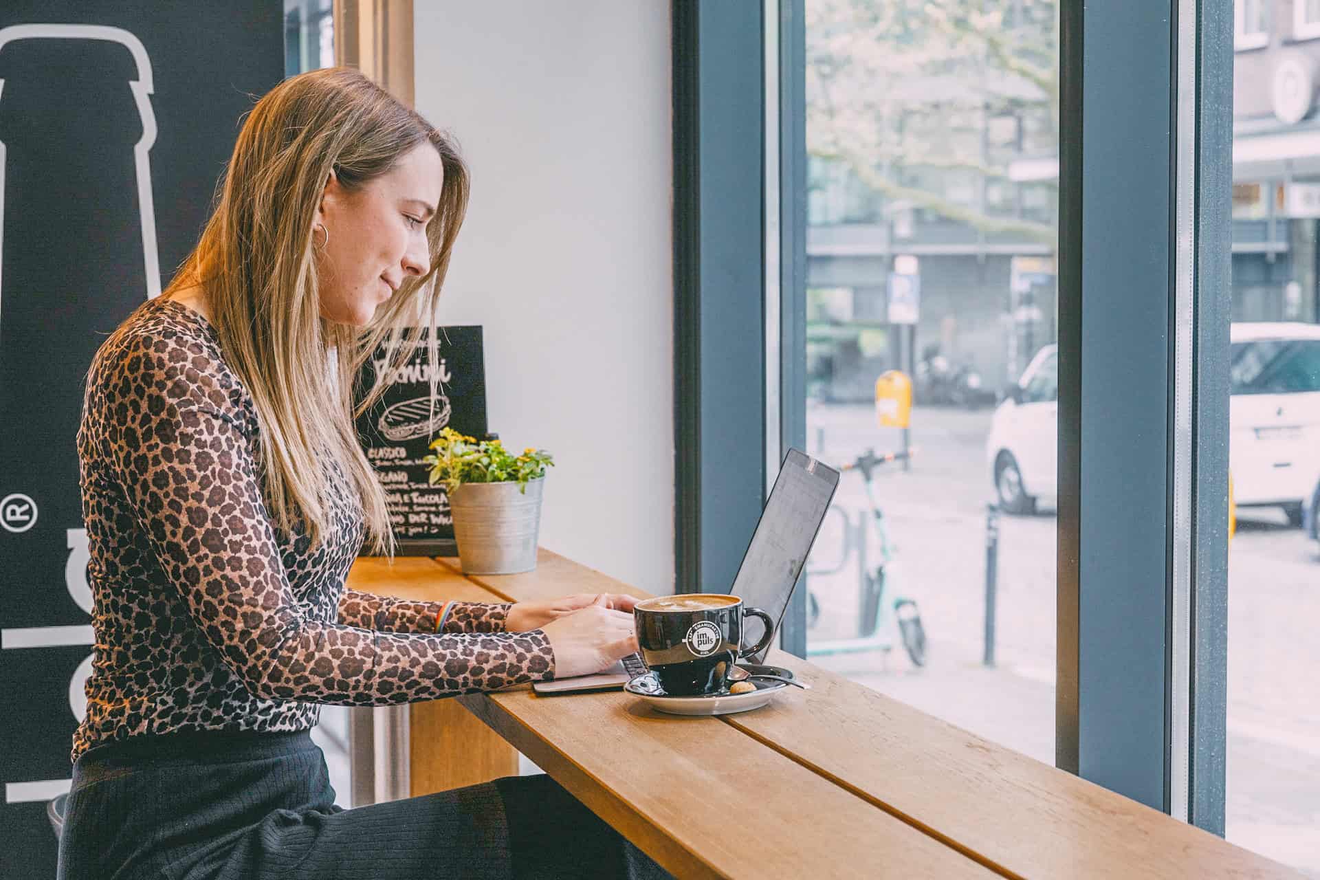 Finja sitzt mit einer Tasse Cappuccino am Tisch und arbeitet im Café am Laptop