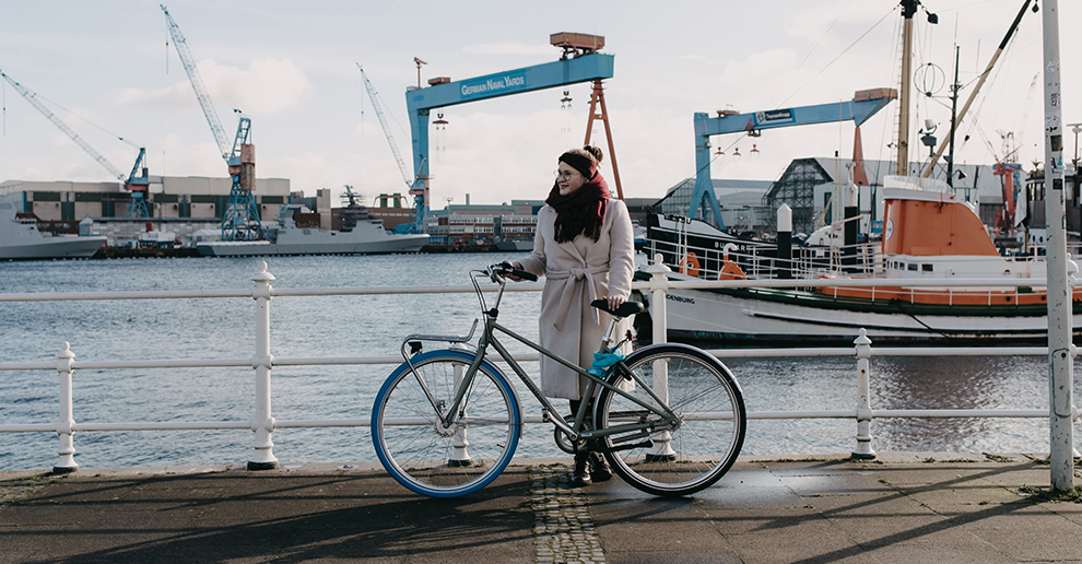 Mädchen mit beigem Mantel und Swapfiets-Fahrrad steht am Wasser