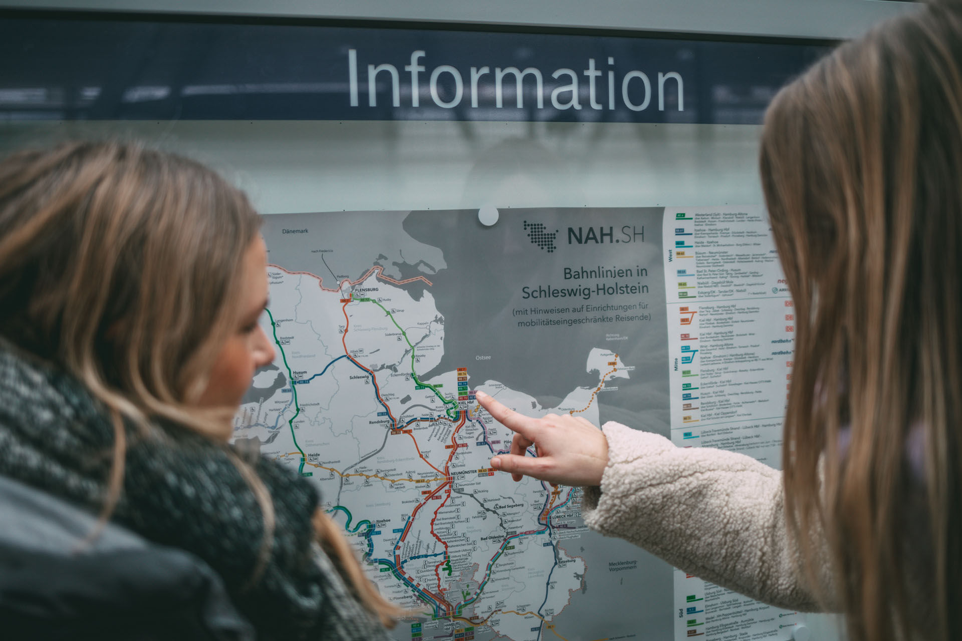 Finja und Sina stehen auf ihrem nachhaltigen Ausflug am Kieler Hauptbahnhof und zeigen auf eine Karte mit Zugverbindungen