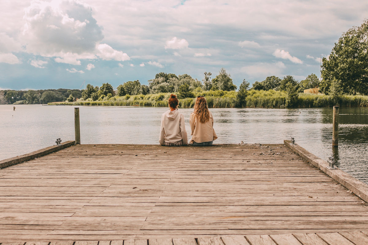Mona und FInja sitzen auf einem Steg und blicken auf einen See