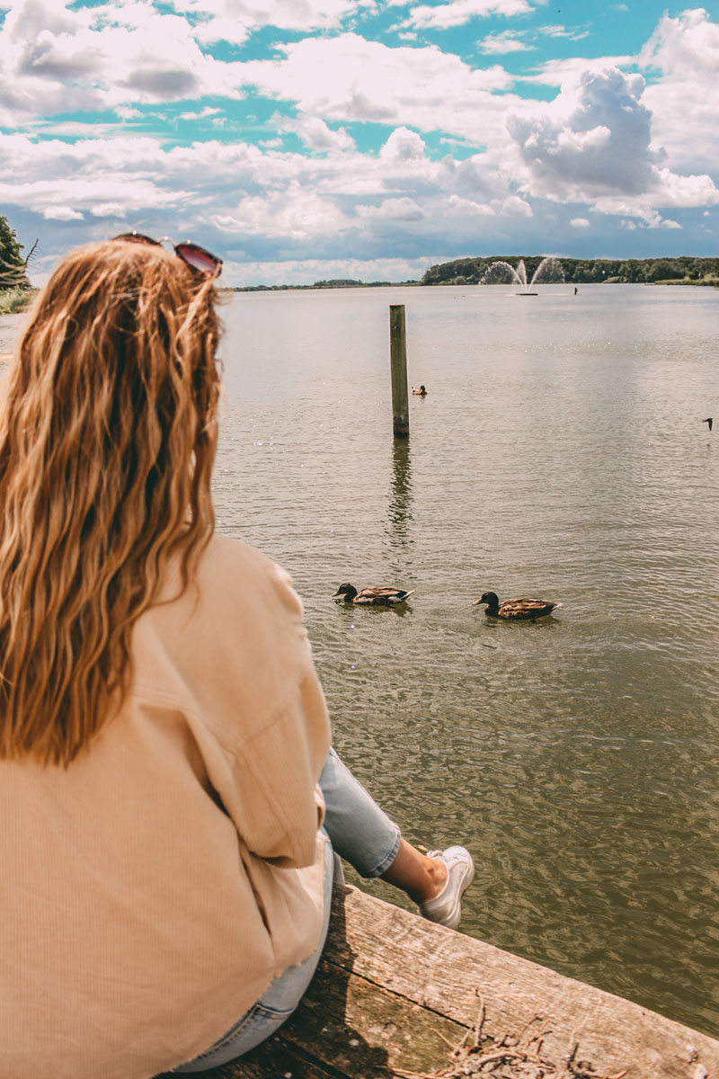 Finja sitzt auf einem Steg und blickt auf einen See. Darin schwimmen zwei Enten 