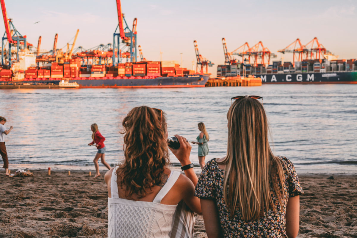 Mona und Finja sitzen am Elbstrand und blicken auf die die Elbe und Containerschiffe