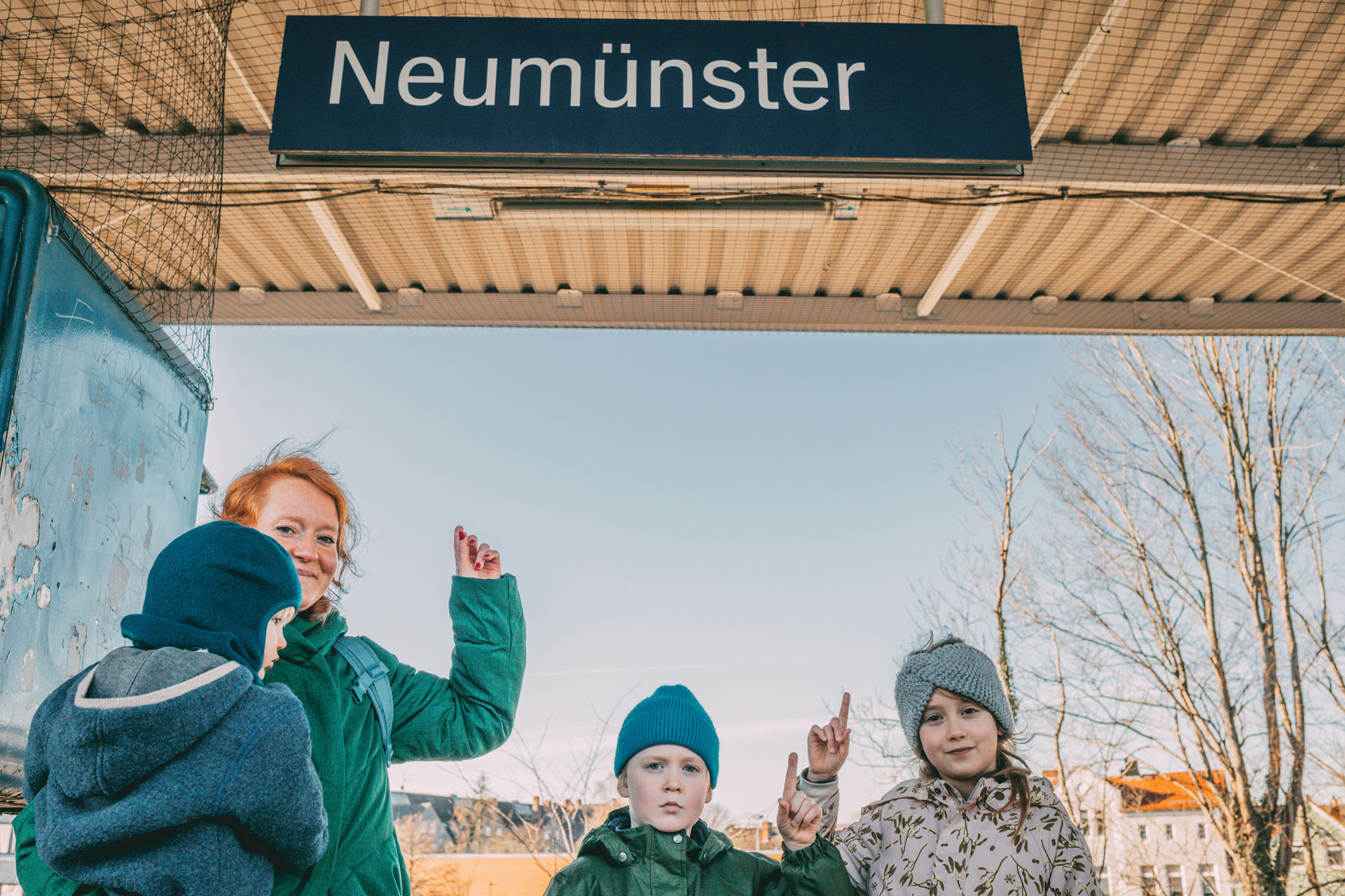 Eine Familie zeigt auf ein Bahnschild mit der Aufschrift „Neumünster“