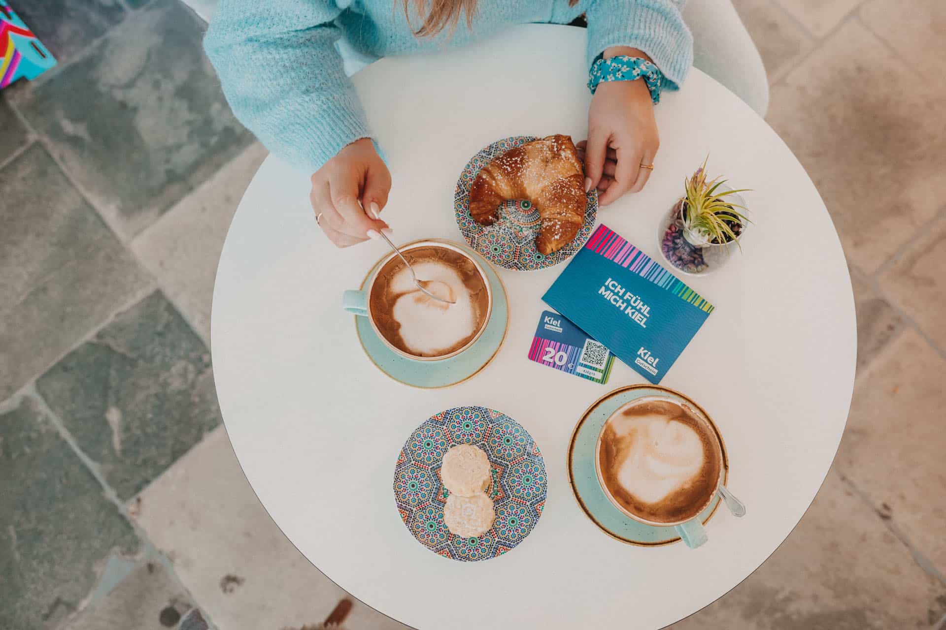 Ein Kaffeetisch von oben. Auf ihm darauf stehen 2 Tassen mit Cappucchino, 2 Teller mit Croissant und Keksen und ein KielGutschein