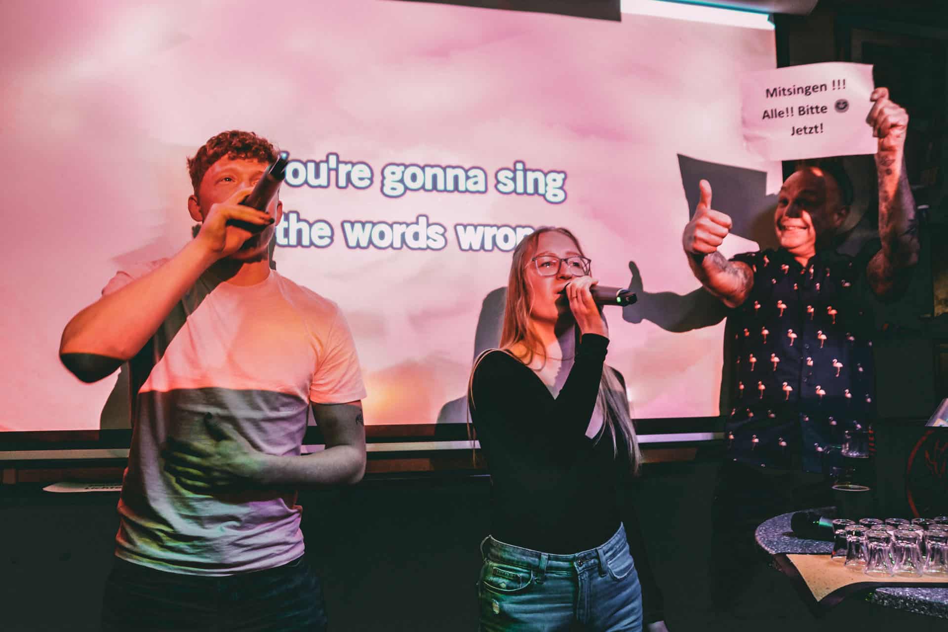 Zwei Personen singen und „Maschine“ hält ein Schild hoch mit der Aufschrift: „Mitsingen! Alle! Bitte! Jetzt!“