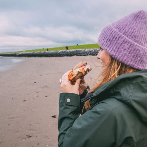 Maya steht am Strand an der Nordsee und isst ein Fischbrötchen