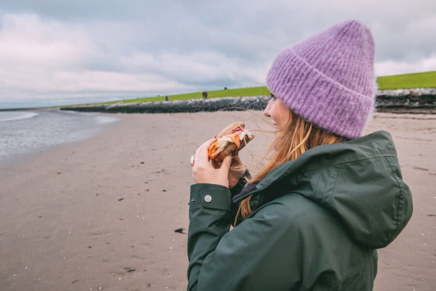 Maya steht am Strand an der Nordsee und isst ein Fischbrötchen