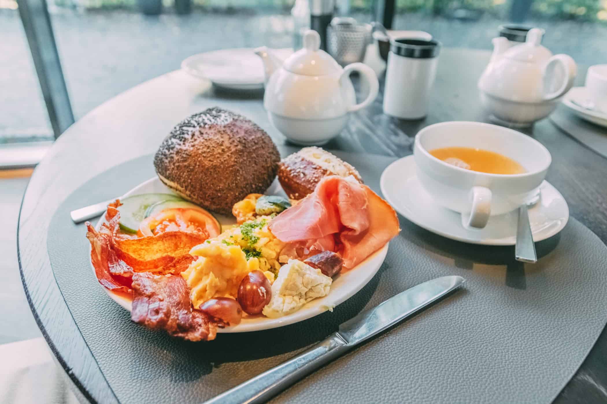 Frühstücksteller mit Brötchen, Bacon, Rührei, Gemüse und Obst. Daneben eine Tasse Tee