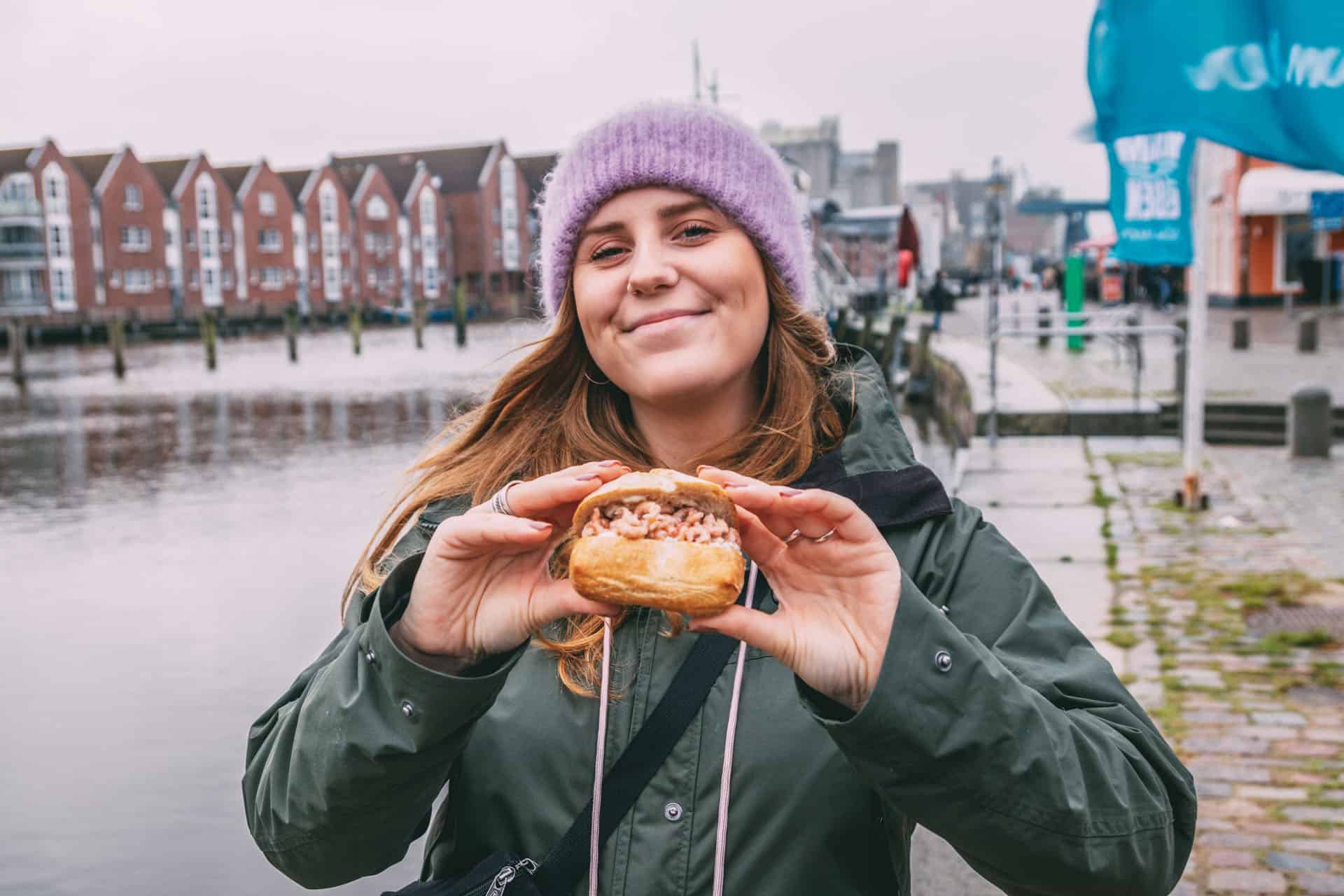 Maya hält mit beiden Händen ein Krabbenbrötchen und lächelt in die Kamera