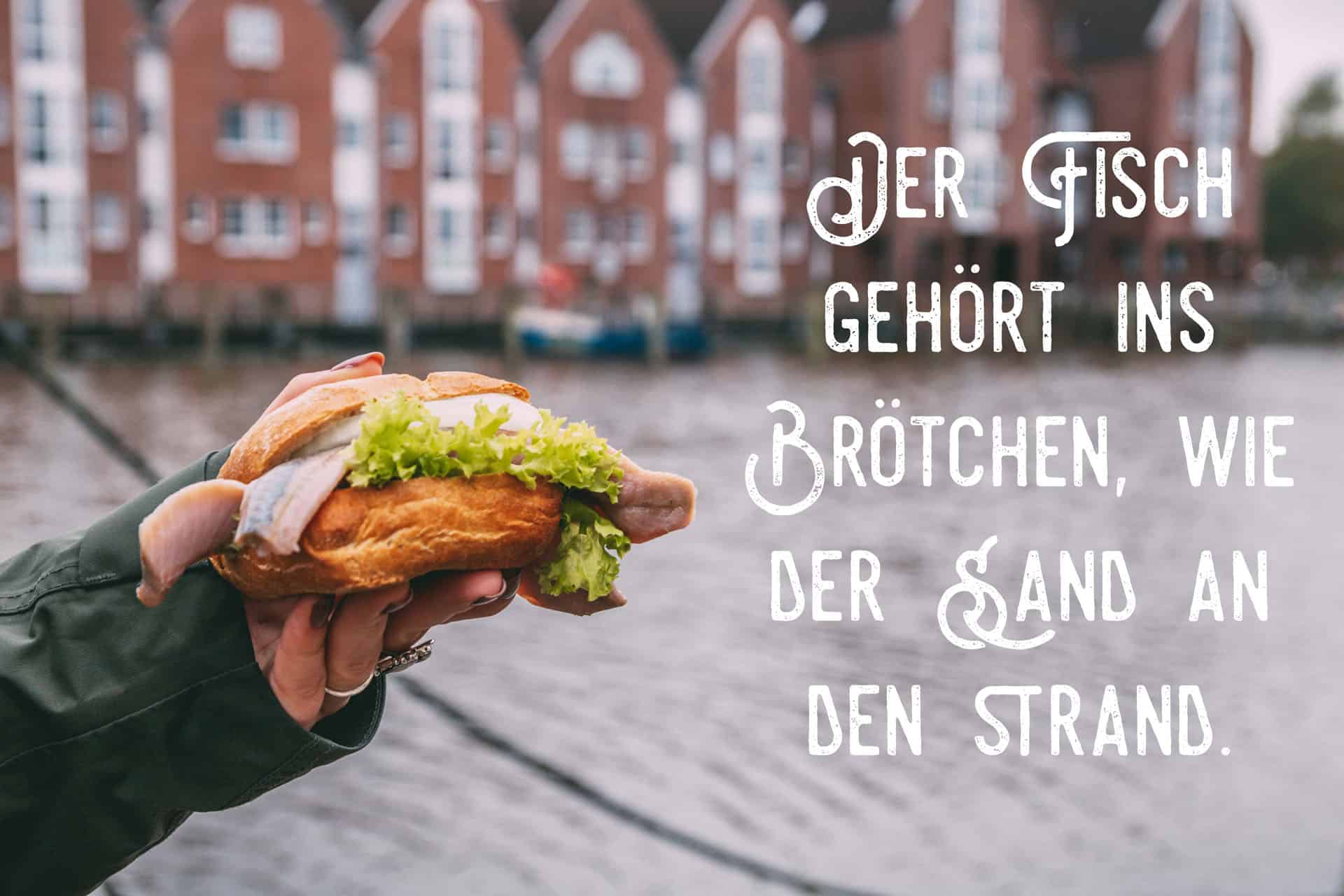 Eine Hand hält ein Fischbrötchen. Im Hintergrund ist Wasser zu sehen. Das Bild trägt die Aufschrift: Der Fisch gehört ins Brötchen, wie der Sand an den Strand