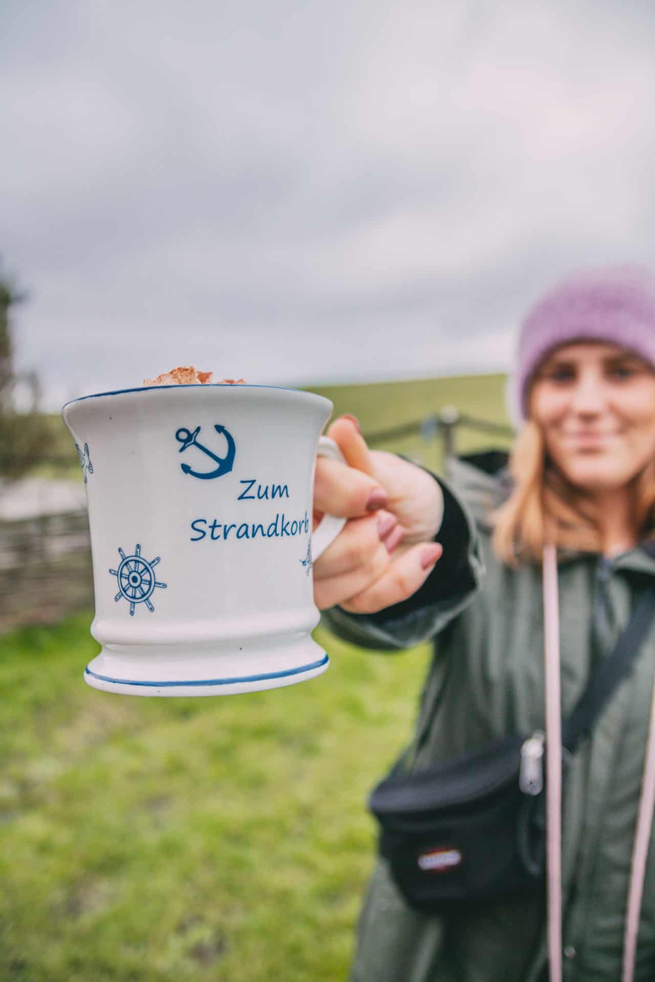 Maya hält eine Tasse Tote Tante in die Kamera. Die Tasse trägt die Aufschrift „Zum Strandkorb“