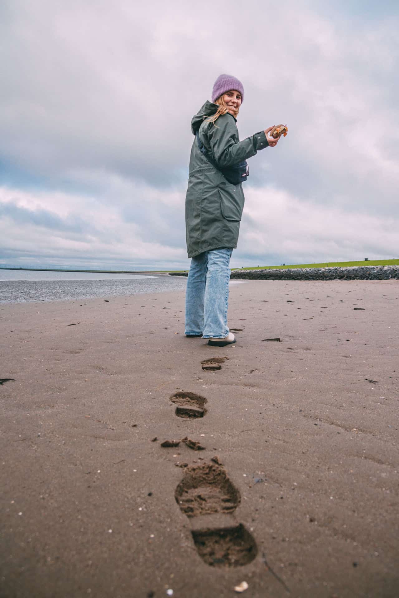 Maya läuft im Sand und hält ein Fischbrötchen. Im Sand sind ihre Fußspuren zu sehen