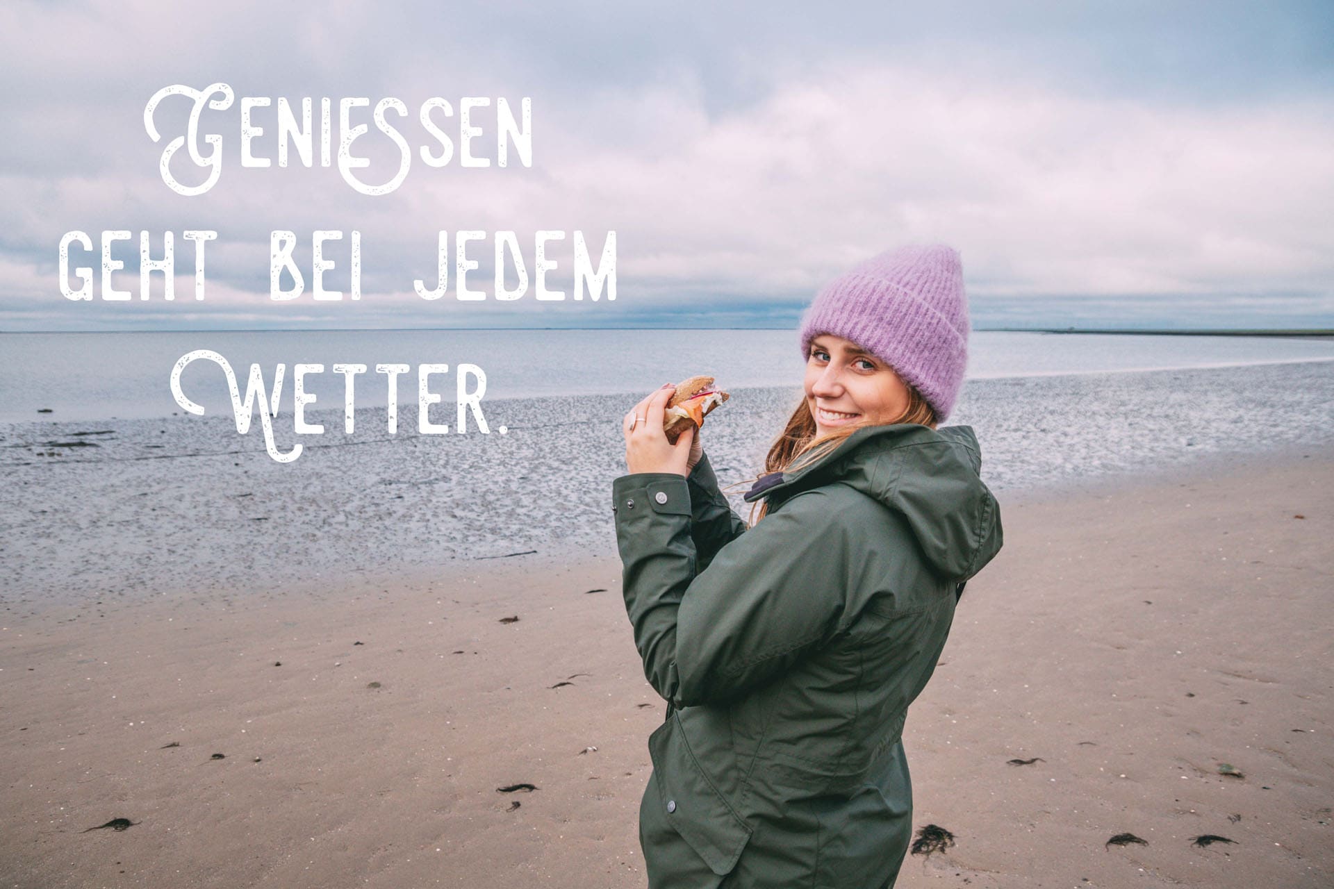 Maya steht am Strand an der Nordsee und hält ein Fischbrötchen in den Händen. Sie trägt eine Regenjacke und Mütze. Dazu die Aufschrift: Genießen geht bei jedem Wetter